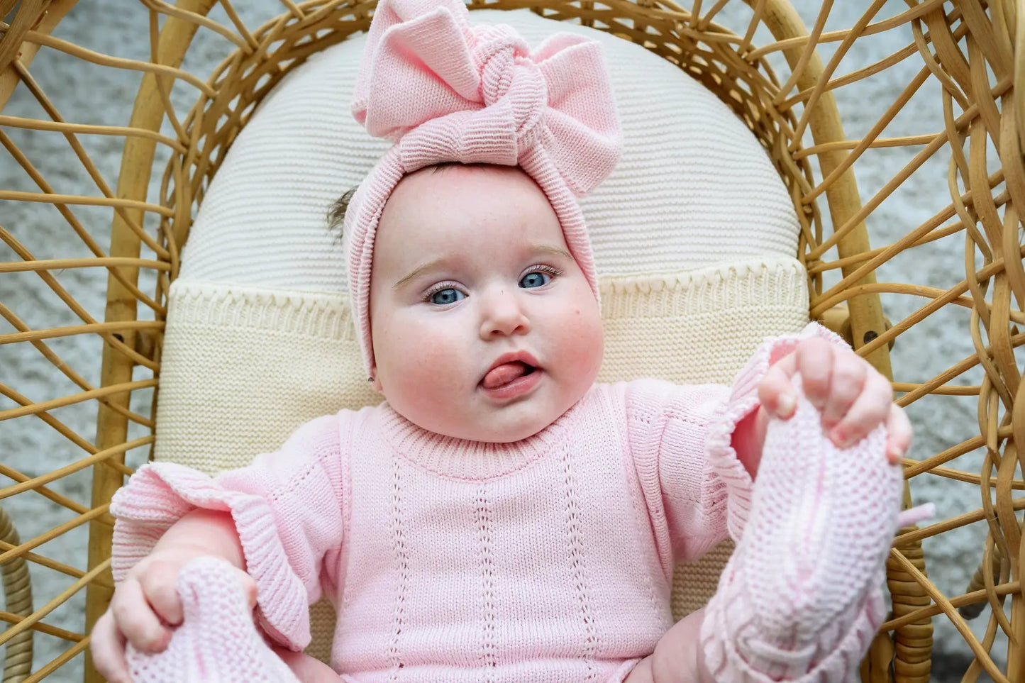 Baby wearing pink knitwear outfit and headband sitting in wicker chair