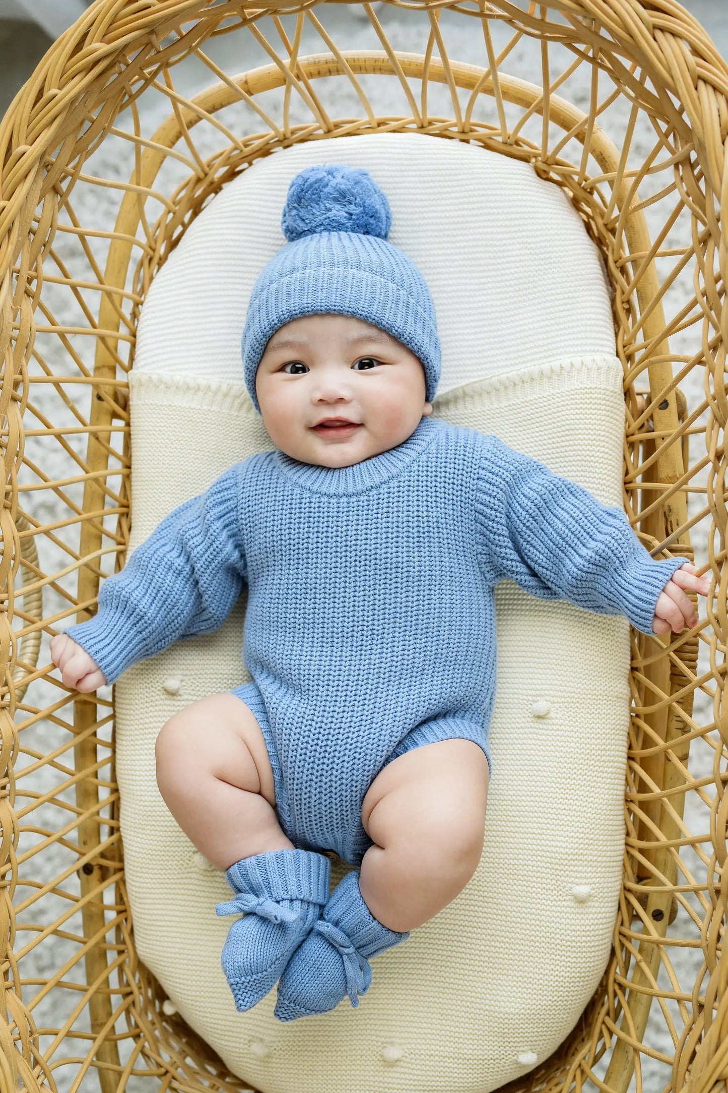 Smiling baby in blue knit romper, beanie, and booties lying in a wicker bassinet