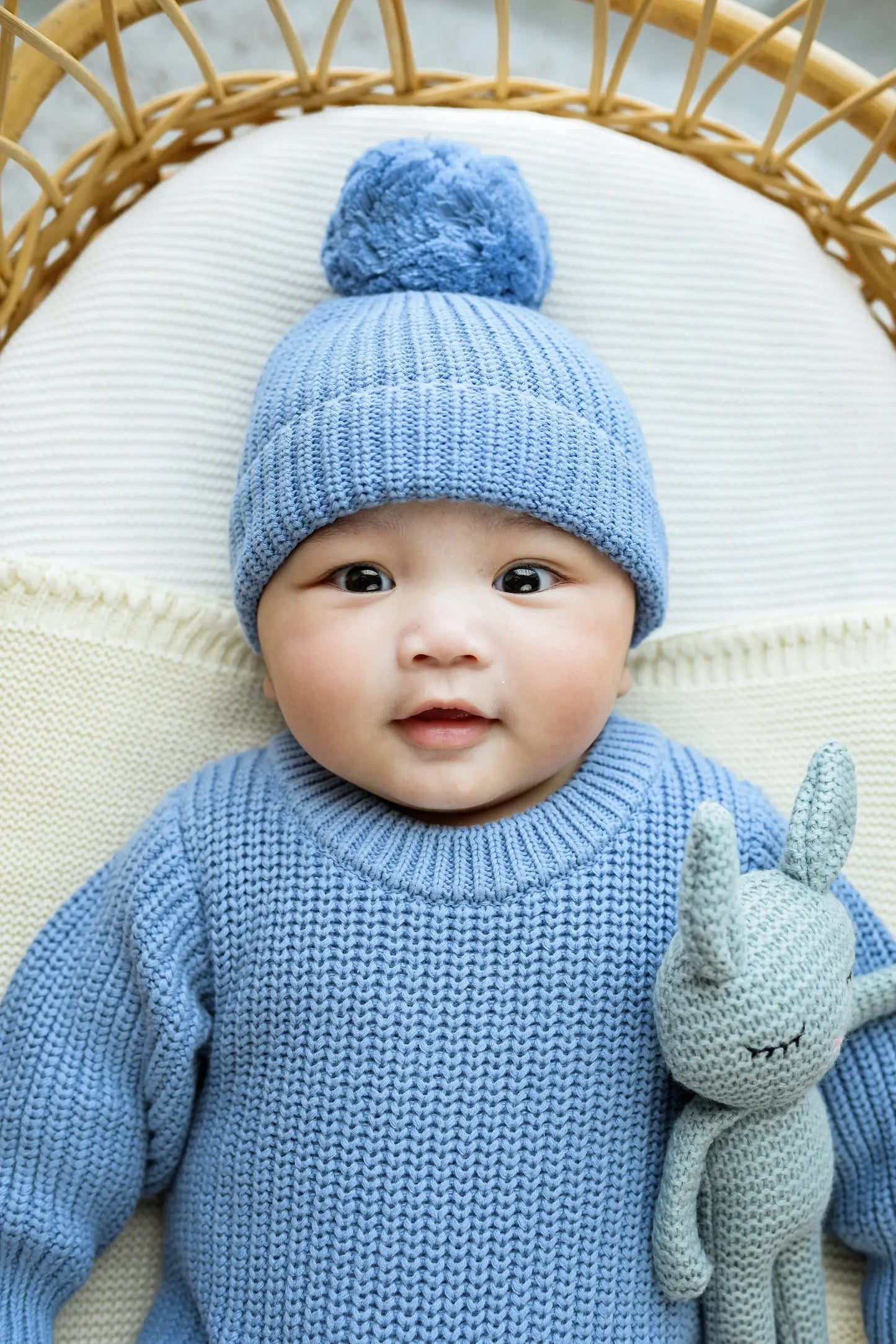 Baby in a blue knit sweater and beanie holding a soft toy, cozy and smiling