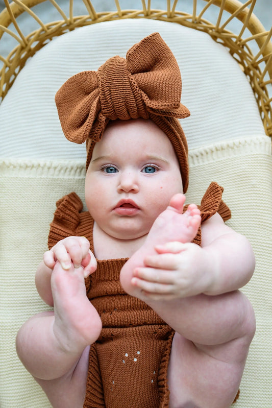 Baby in brown knit romper and matching bow headband sitting on cream blanket