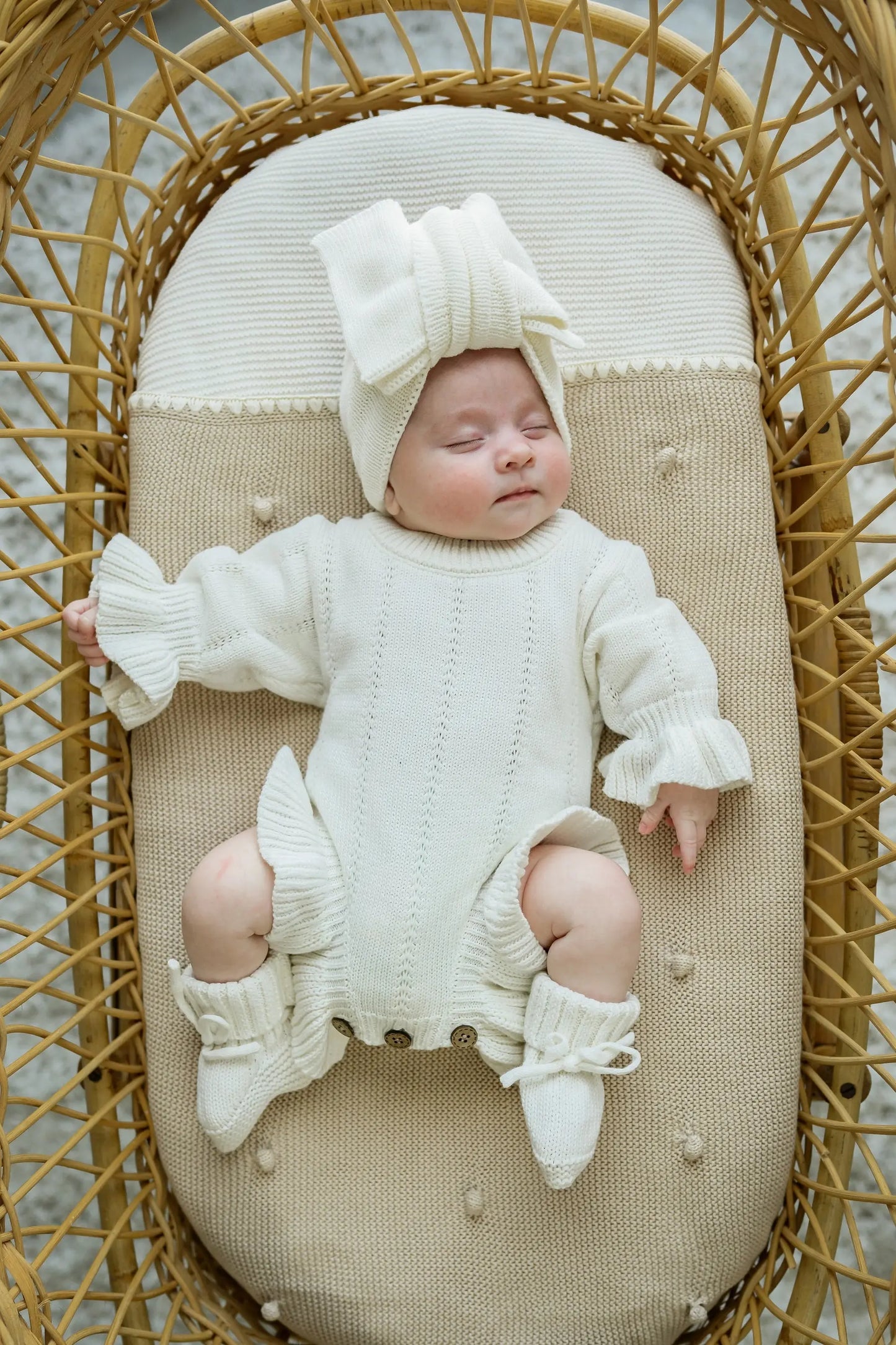 Baby wearing cream knit romper, booties, and headband, lying in a woven bassinet