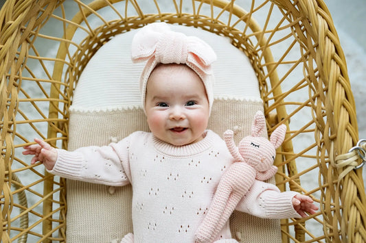 Smiling baby in pink knitwear and headband holding a knit bunny in a wicker bassinet