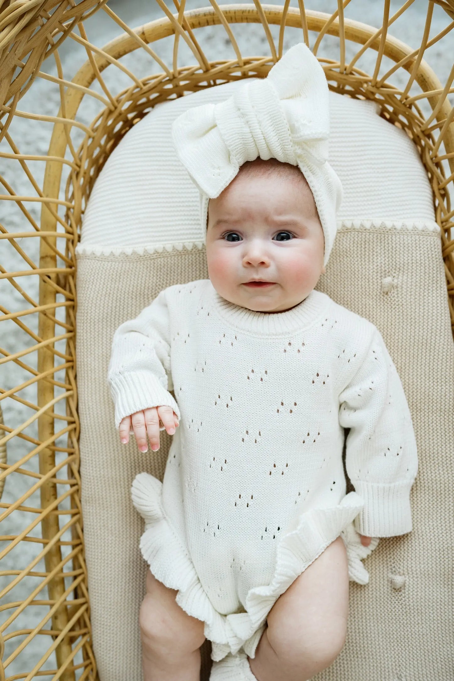 Baby in cream knit romper and bow headband lying on rattan bassinet