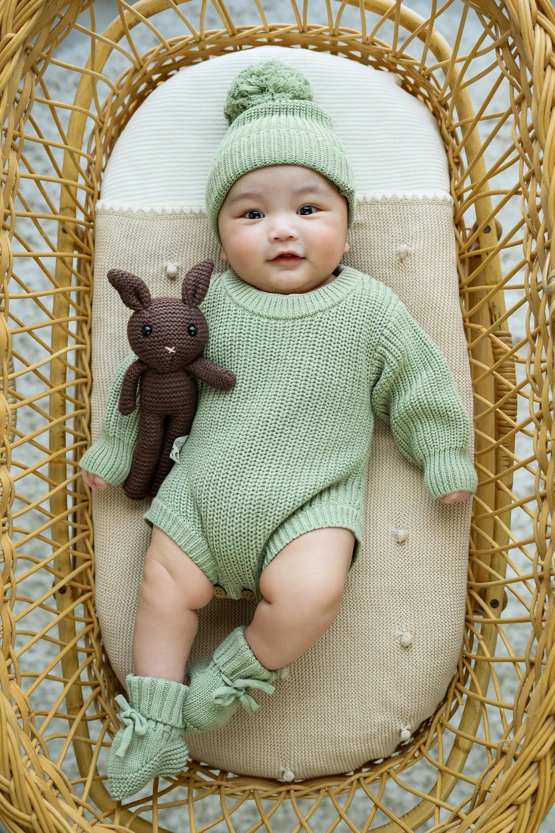 Baby in sage green knit romper, hat, and booties, lying in basket with brown bunny toy