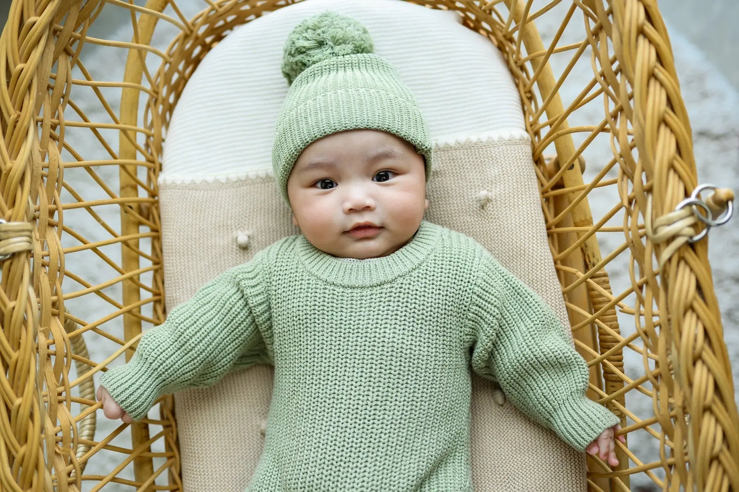 Baby in green knit sweater and beanie sitting in rattan chair, cozy toddler clothing