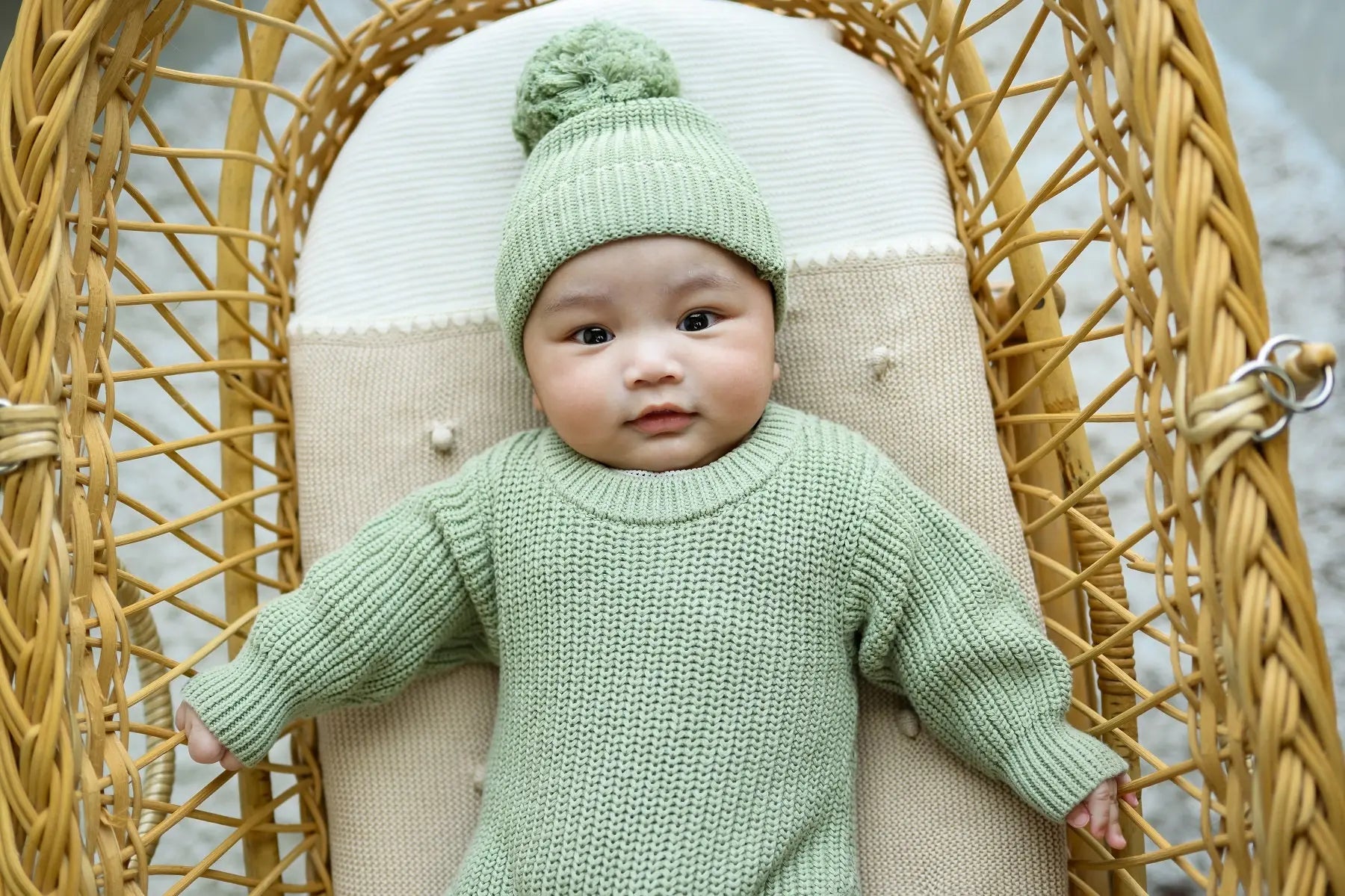 Baby in green knit sweater and beanie sitting in rattan chair, cozy toddler clothing