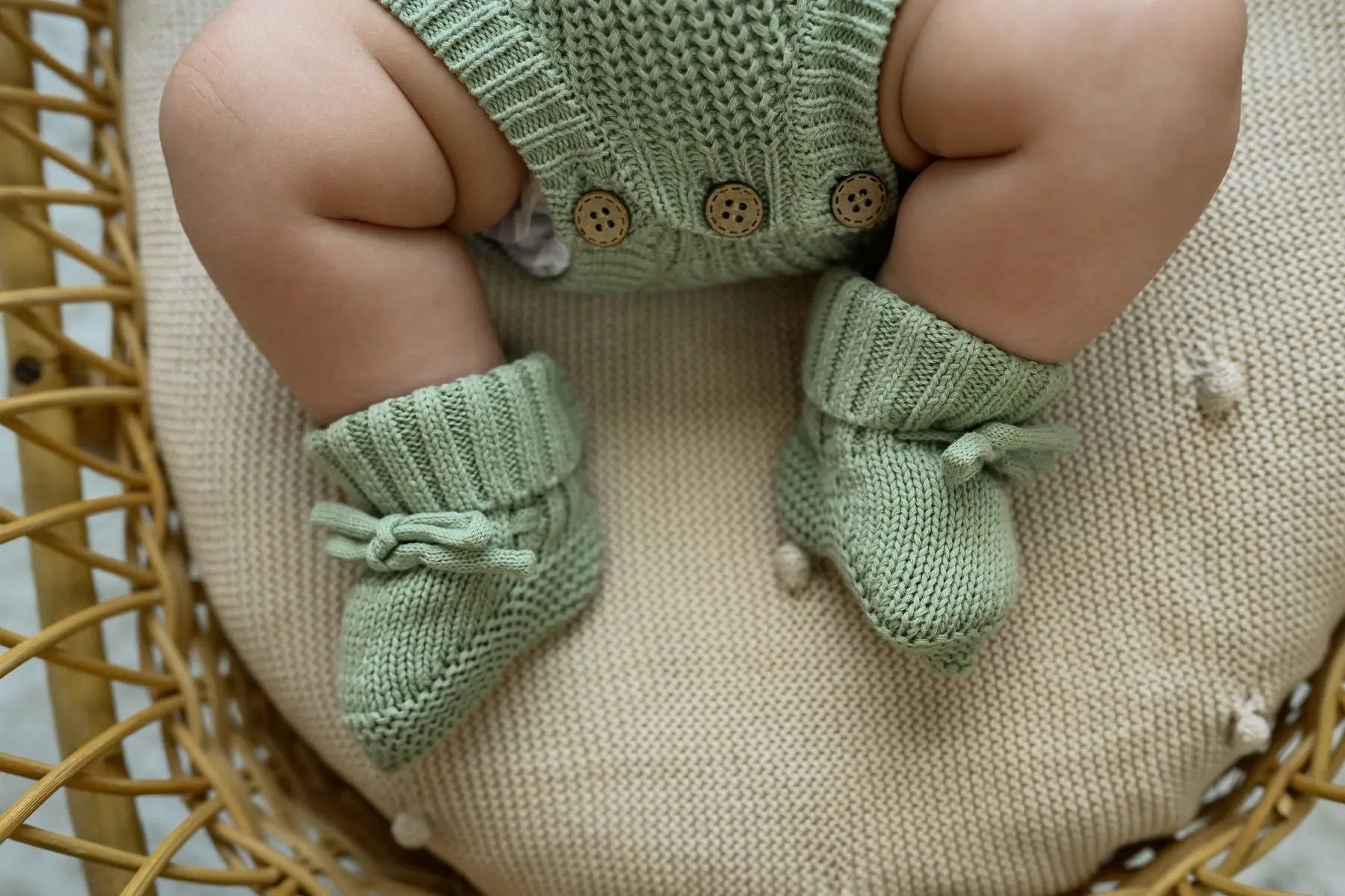 Baby wearing sage green knit romper and booties on a textured cushion