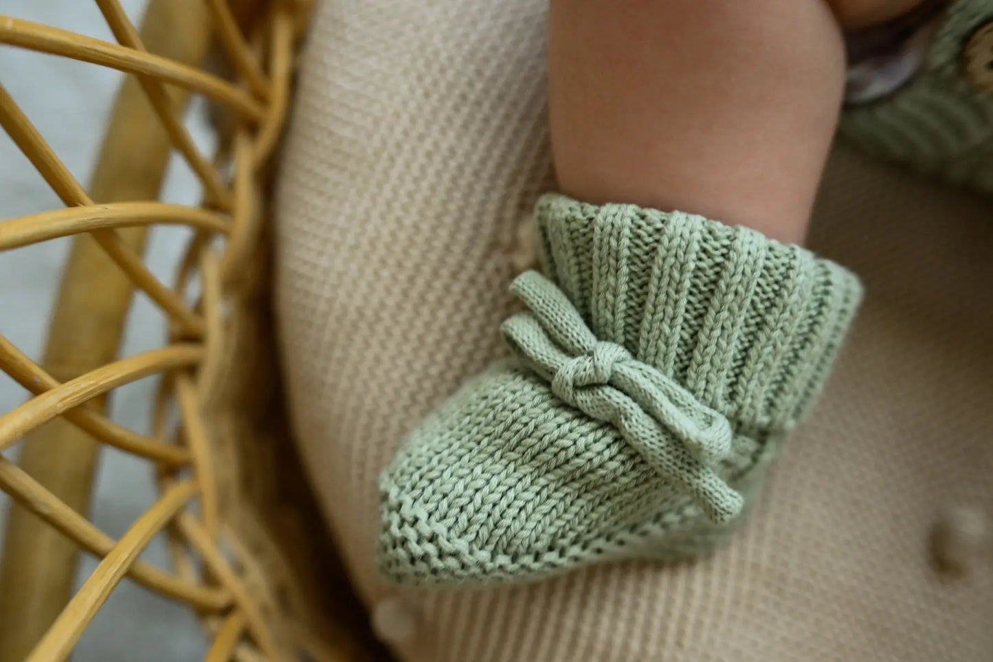 Close-up of baby wearing soft green knit booties with bow detail on a textured cushion.