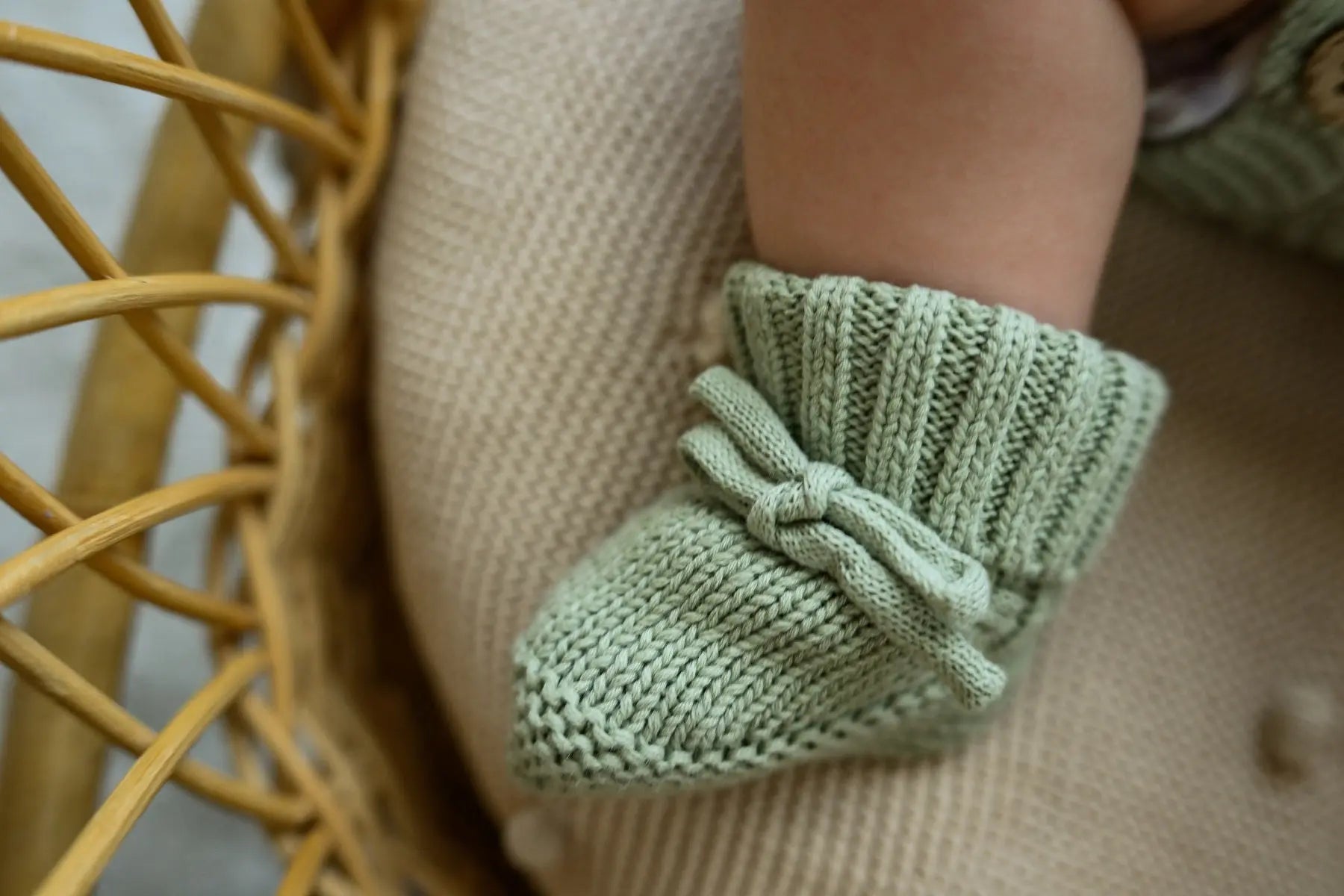 Close-up of baby wearing soft green knit booties with bow detail on a textured cushion.