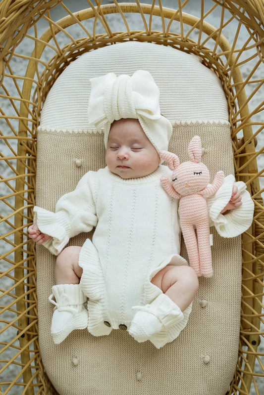 Baby sleeping in a wicker bassinet, wearing cream knit romper and headband, holding pink plush toy
