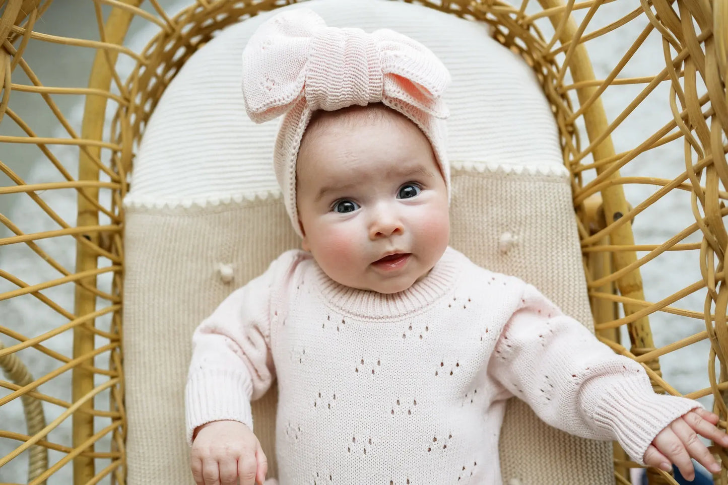 Baby in soft pink knit sweater and matching bow headband sitting in a rattan chair