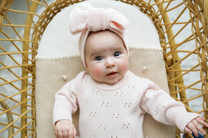 Baby in soft pink knit sweater and matching bow headband sitting in a rattan chair