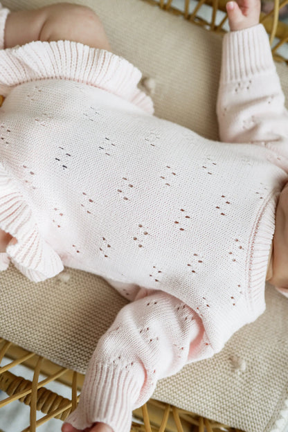 Baby in soft pink knit jumpsuit with delicate eyelet patterns, lying on beige blanket