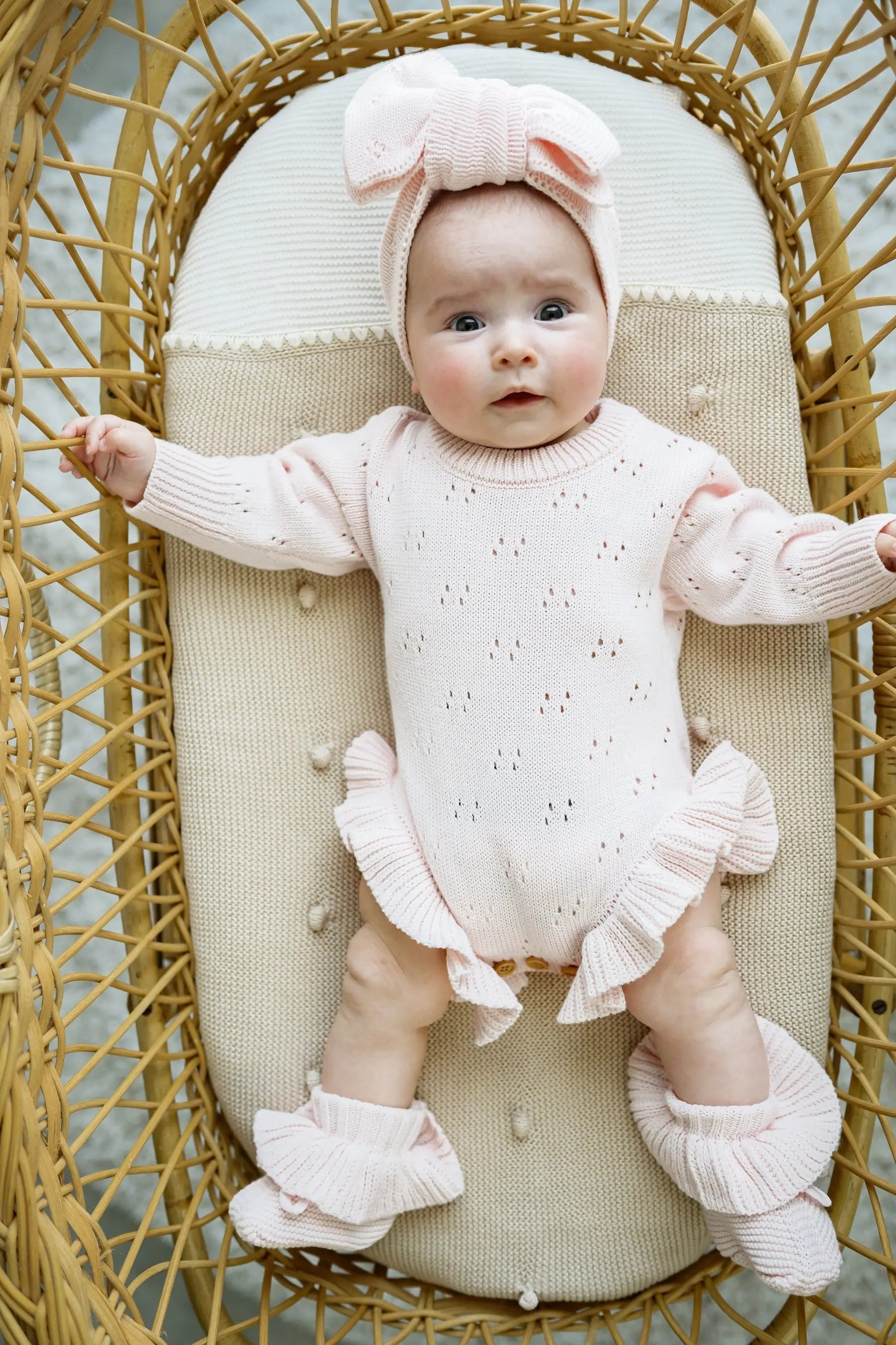 Baby in pale pink knit romper with ruffles and matching headband, lying in wicker bassinet