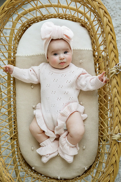 Baby in a pink knit ruffle romper and headband lying in a wicker bassinet