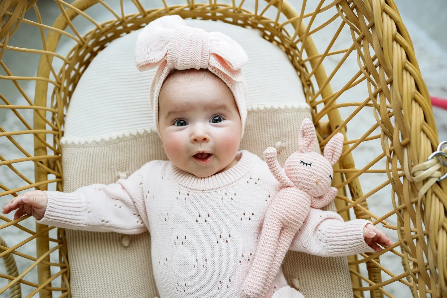 Baby in a pale pink knit outfit and bow headband, holding a pink knit bunny in a wicker chair