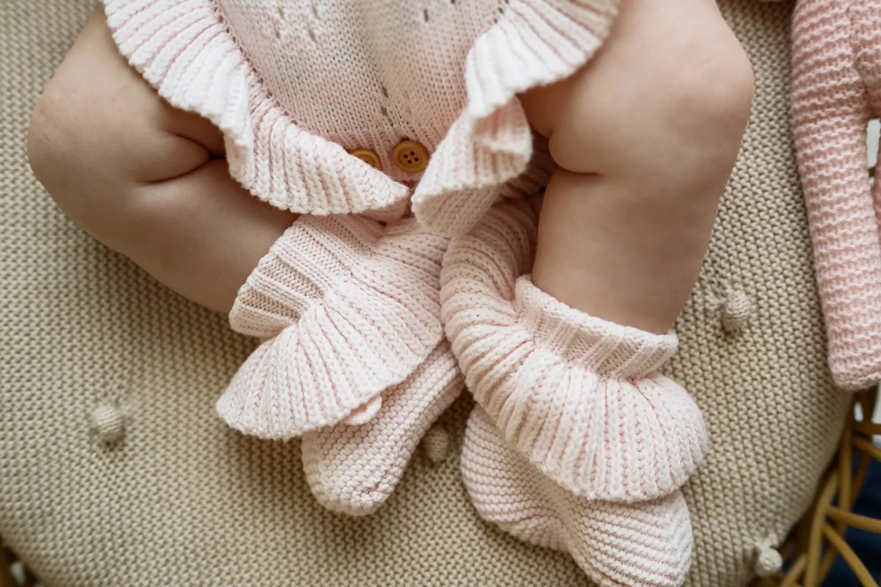 Baby wearing soft pink knit booties and romper sitting on a textured beige cushion